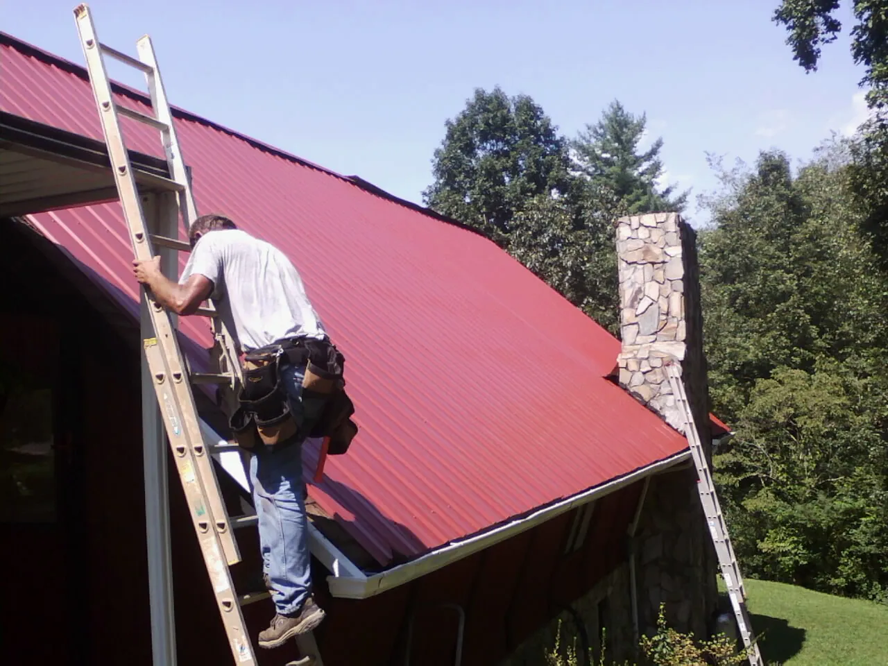 Roofer on a ladder beside a red metal roof and stone chimney — Soffit & Fascia Repair by Roofing Brothers in Brownstown